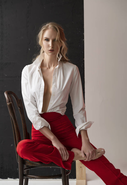 A beautiful blonde curly girl wearing a red pants and a blouse, unbuttoned on her braless big breasts, poses sitting on the chair in the studio. Advertising, fashionable, commercial, design. - Fotó, kép