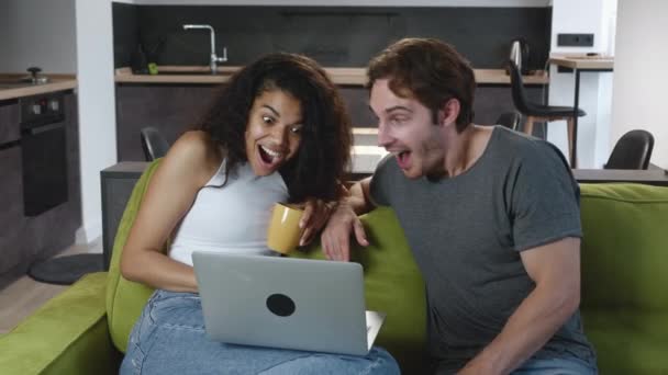 Excited happy multiracial couple looking at screen of laptop feel winners surprised by lottery betting winning bid. Lovely boyfriend and girlfriend celebrating internet lottery victory prize good news - Footage, Video