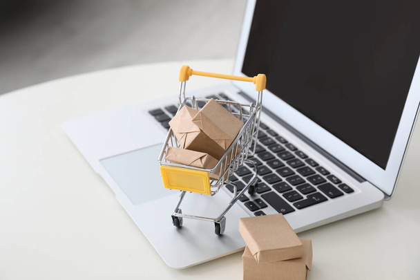 Internet shopping. Laptop and small cart with boxes on white table indoors - Foto, imagen