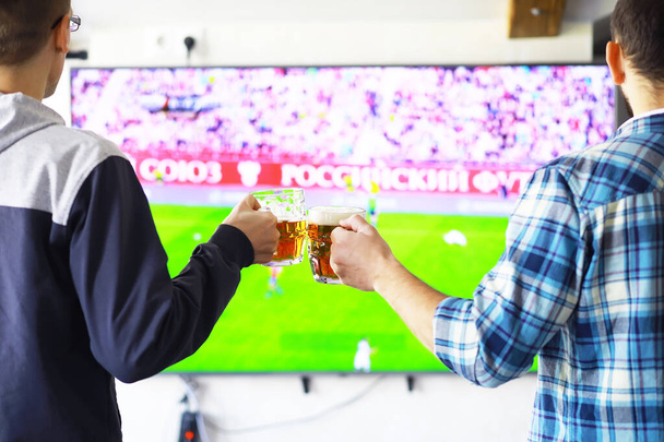Hands horizontal holding lager beer glass and clinking on background of football game. Sport fans cheer up. Friends leisure lifestyle concept. - Photo, Image