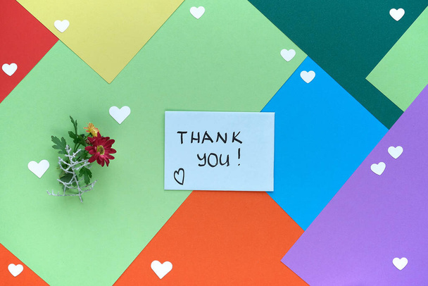 International Thank you day. Greeting card with female hand holding envelope with heart shape and flowers. Thank you handwriting on card. Concept flat lay on multicolor layered paper background. - 写真・画像