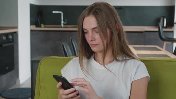 Ανήσυχη γυναίκα διαβάζει άσχημα νέα στο μήνυμα στο smartphone της, ενώ κάθεται στον καναπέ στο σπίτι. Σοκαρισμένη νεαρή γυναίκα κρατώντας το κινητό τηλέφωνο στα χέρια στο σαλόνι - Πλάνα, βίντεο