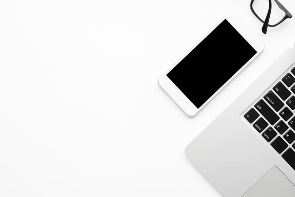 Белый смартфон с черным пустым макетом экрана находится на верхней части белого стола офиса стол с ноутбуком. Вид сверху с копировальным пространством, плоская кладка. - Фото, изображение
