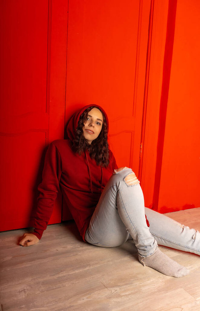 Young woman in hood sitting on floor. Curly brunette poses near red wall - Foto, imagen