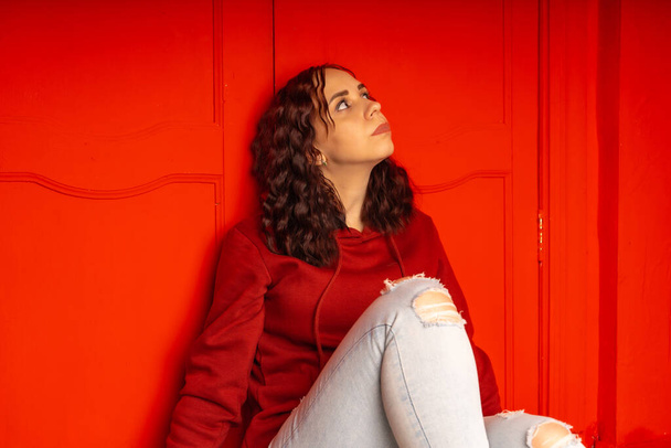 Young woman sitting on floor. Curly brunette poses near red wall - Фото, зображення