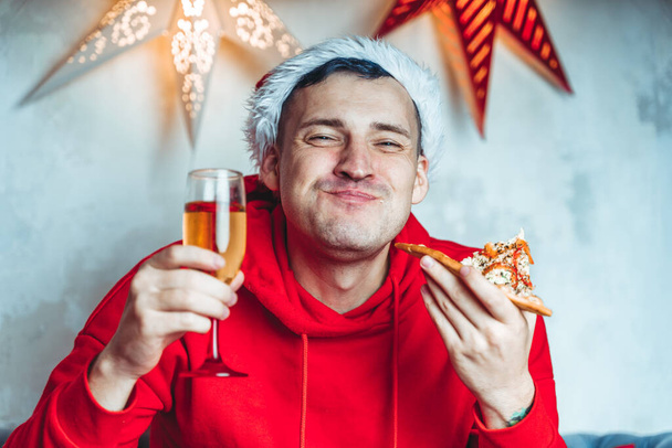 Young man in santa hat drinks champagne and eats pizza, sitting on sofa in room. Happy male resting with food and alcohol. Concept of Christmas celebration at home - Photo, Image