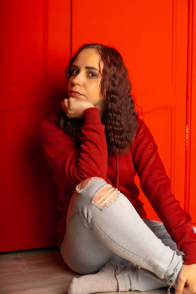 Young woman sitting on floor. Curly brunette poses near red wall - Photo, image