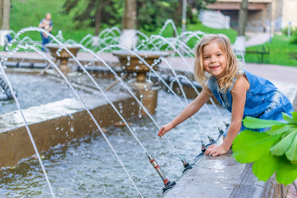 Μικρή όμορφη ξανθιά κοπέλα παίζεται σε ένα σιντριβάνι σε ένα πάρκο της πόλης το καλοκαίρι. - Φωτογραφία, εικόνα