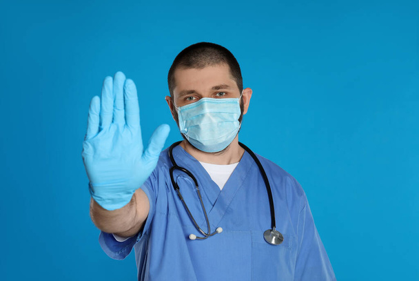 Doctor in protective mask showing stop gesture on light blue background. Prevent spreading of coronavirus - Foto, Bild