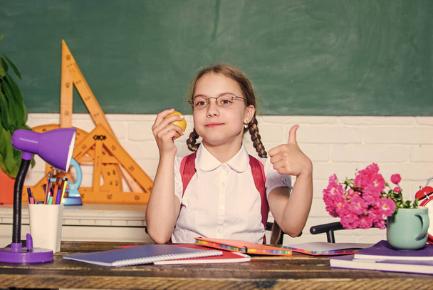 Schoolgirl sit desk chalkboard background. Kid student in school. Girl small child eating apple snack. School break. Relax between lessons. School life concept. Healthy lifestyle. Modern childhood - Fotografie, Obrázek