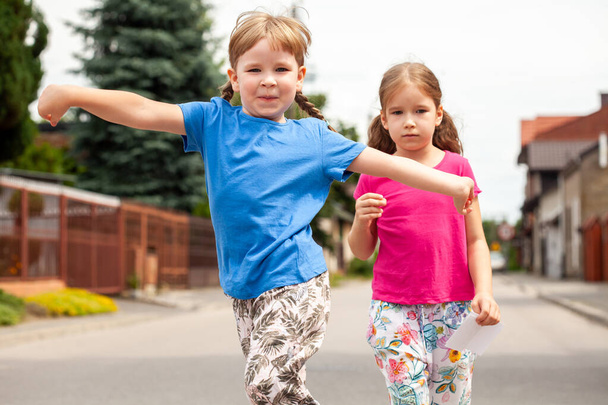 2つの幸せな小さな子供たち、姉妹、兄弟、女の子が喜んで腕を広げてカメラに向かってジャンプ歩く。子供の家族の散歩、町のエリア、自然のライフスタイルシーン、屋外のシンプルな肖像画 - 写真・画像