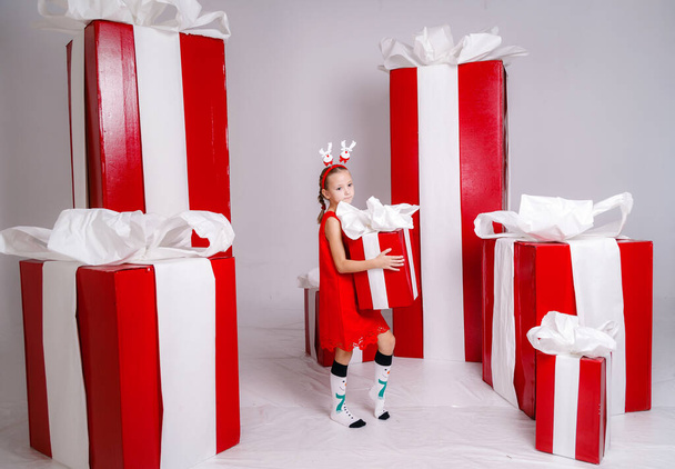 χαρούμενο αστείο κορίτσι παιδί με κοστούμι Χριστούγεννα με μεγάλα κόκκινα δώρα στο παρασκήνιο. - Φωτογραφία, εικόνα