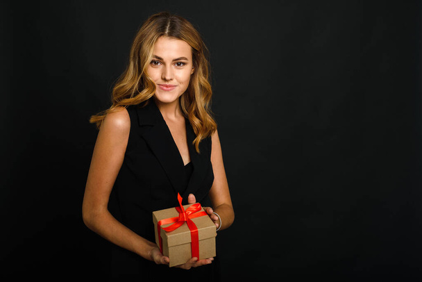 A beautiful young woman on a black background holding a Christmas present with a red ribbon. The happiness of receiving and giving gifts for the new year - Photo, Image