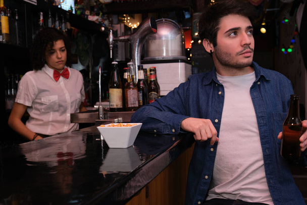Fiatal fehér férfi egy sörösüveggel a kezében TV-t néz, focimeccset egy bárban. Pincérnő a háttérben. - Fotó, kép