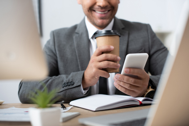 vista ritagliata di un uomo d'affari sorridente che chiacchiera sullo smartphone mentre tiene il caffè per andare in primo piano sfocato - Foto, immagini
