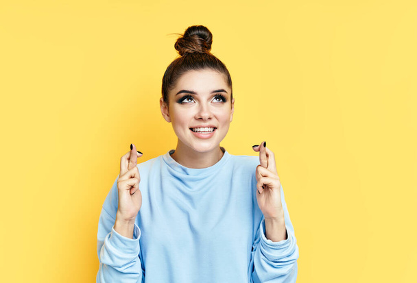Young beautiful woman crossing her fingers and wishing for good luck over yellow background. - Photo, Image