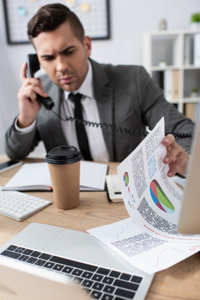 businessman talking on landline phone while looking at papers with infographics, blurred background - Photo, Image