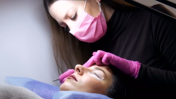 Face of a young girl before a modern eyelash lamination procedure in a professional beauty salon. The master applies special glue before the eyelash curling procedure in pink rubber gloves and a mask - Footage, Video
