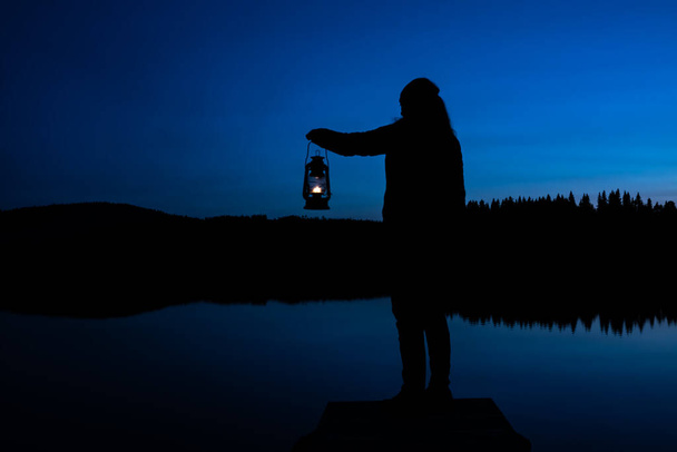 Silhouette of a woman looking into the last light of the day by a lake in the wild, holding a lit vintage kerosene lamp. . High quality photo - Photo, Image