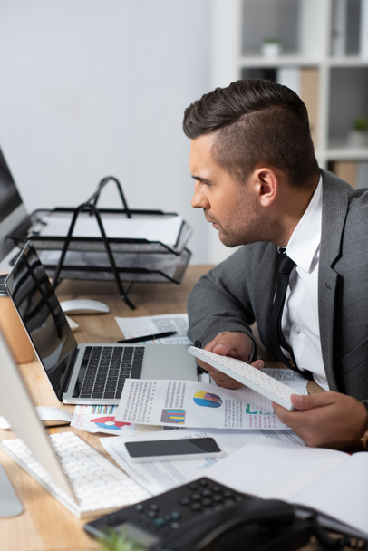 homem de negócios concentrado olhando para laptop perto de papéis com gráficos e gráficos, foreground borrado - Foto, Imagem