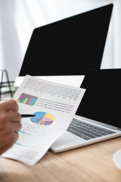 обрезанный вид трейдера, указывающий ручкой на инфографику возле ноутбука и монитор с пустым экраном, размытый передний план - Фото, изображение