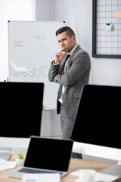 thoughtful trader standing at flipchart with graphs near monitors with blank screen, blurred foreground - Photo, Image