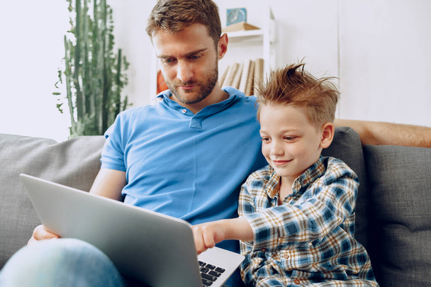 Father and son sitting on couch and watching something on laptop - Zdjęcie, obraz
