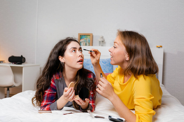 İki genç kadın yatağa uzanıp dekoratif kozmetik ürünleri yapıyorlar. Bir kadın başka bir kadının kirpiklerini maskarayla boyuyor. LGBT ilişkileri ve makyaj kavramı. - Fotoğraf, Görsel