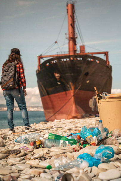 Eine Frau mit einem Rucksack auf dem Rücken steht auf dem Hintergrund des Meeres und des Schiffes in Großaufnahme. Im Vordergrund ein Müllcontainer und verstreute Plastikflaschen. Vertikal. - Foto, Bild