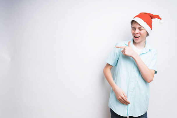 A mid shot of a funny teenage boy in a Santa Claus hat on a white background. He's checking something out and pointing to the side - Valokuva, kuva