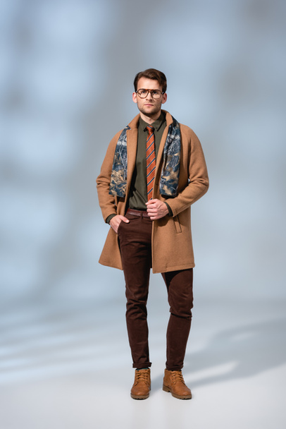 full length of trendy man in winter coat standing with hand in pocket on grey - Fotografie, Obrázek