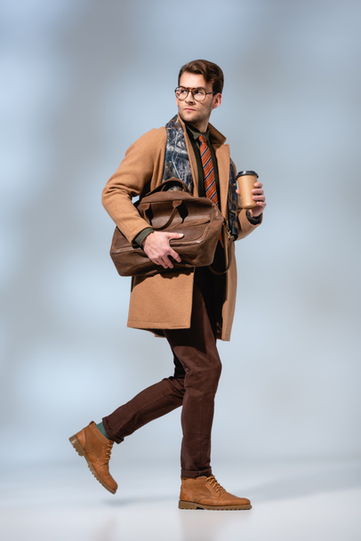 full length of stylish man in winter coat holding paper cup and leather briefcase while walking on grey  - Fotó, kép