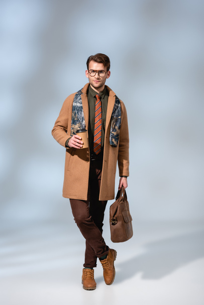 полная длина стильный человек в зимнем пальто держа бумажную чашку и кожаный портфель, стоя со скрещенными ногами на сером  - Фото, изображение