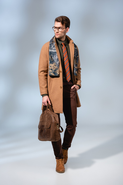 volle Länge der stilvolle Mann im Wintermantel hält Lederaktentasche und steht mit der Hand in der Tasche auf grau  - Foto, Bild