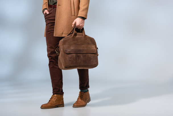 częściowy widok stylowego mężczyzny trzymającego skórzaną walizkę i stojącego z ręką w kieszeni na szarym  - Zdjęcie, obraz