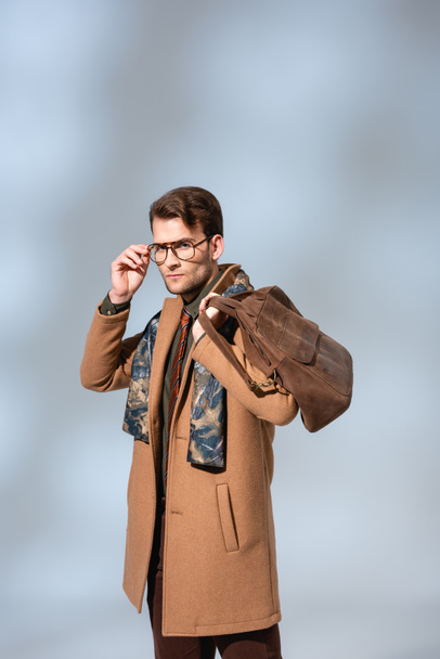 stylish man in winter coat adjusting glasses while holding leather briefcase on grey  - Fotoğraf, Görsel