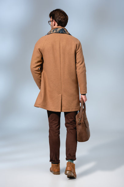 vista posteriore di uomo alla moda in cappotto invernale con valigetta in pelle su grigio  - Foto, immagini