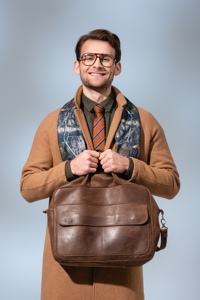 cheerful man in winter coat holding leather briefcase on grey  - Fotó, kép
