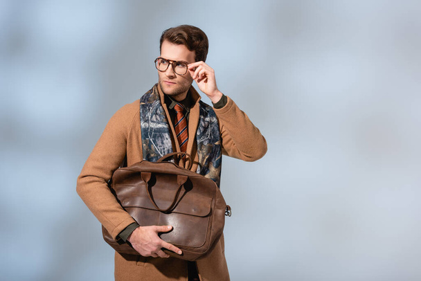 stylish man in winter coat adjusting glasses while holding brown briefcase on grey - Fotó, kép