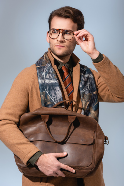 stylish man in winter coat adjusting glasses while holding brown bag on grey - Zdjęcie, obraz