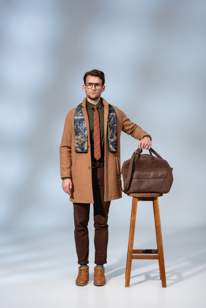 full length of stylish man in winter coat and glasses standing near wooden chair and holding briefcase on grey - Foto, imagen