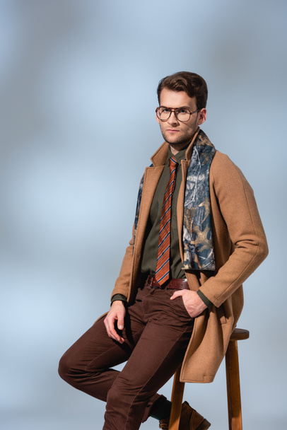 stylish man in winter outfit and glasses sitting on wooden chair with hand in pocket on grey - Fotó, kép