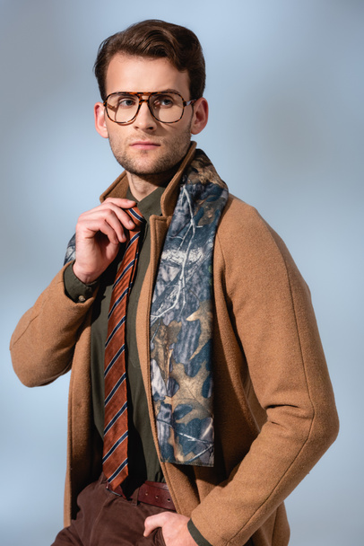 trendy man in winter coat and glasses adjusting tie on grey - Foto, imagen