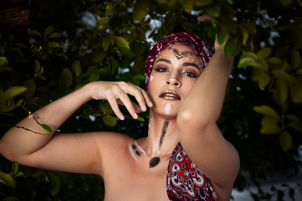 Chica con la cara pintada como habitante nativa de África o del Amazonas, con una bufanda en la cabeza, entre la vegetación, hermosa aborigen - Foto, imagen