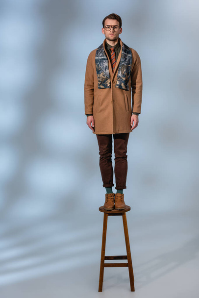 full length of stylish man in winter outfit and glasses standing on wooden stool on grey - 写真・画像