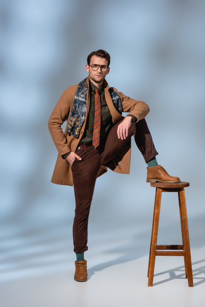 full length of stylish man in winter outfit and glasses posing while leaning on wooden stool on grey - Fotografie, Obrázek