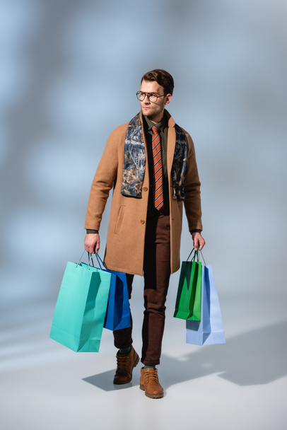 full length of trendy customer in glasses holding paper bags on grey - Fotó, kép