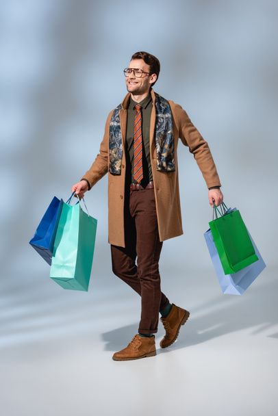 full length of smiling customer in glasses holding paper bags on grey - Foto, immagini