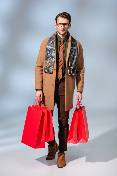 full length of stylish customer in glasses holding paper bags on grey - Photo, image