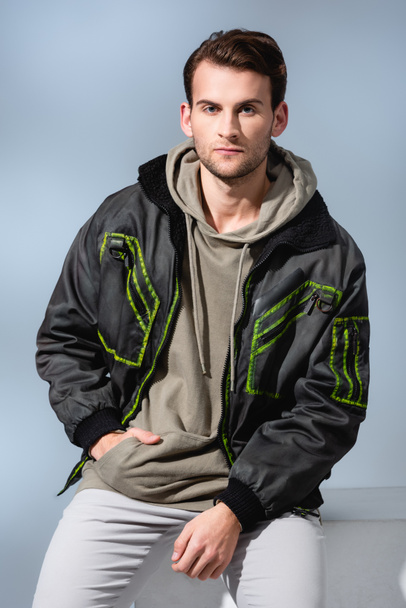 trendy man in hoodie and parka posing while sitting on white cube isolated on grey  - Fotó, kép